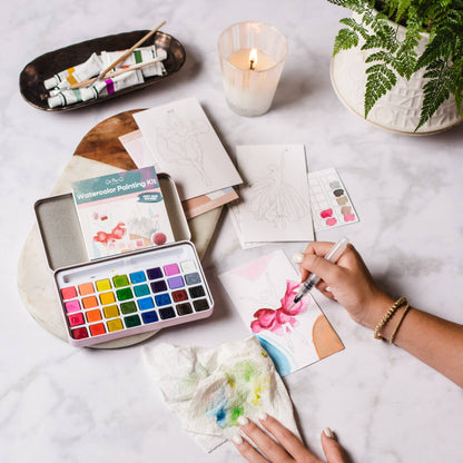 Person painting with watercolors on a table with art supplies and a candle.