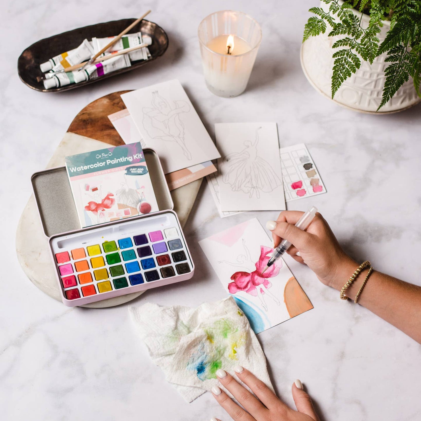 Person painting with watercolors on a table with art supplies and a candle.