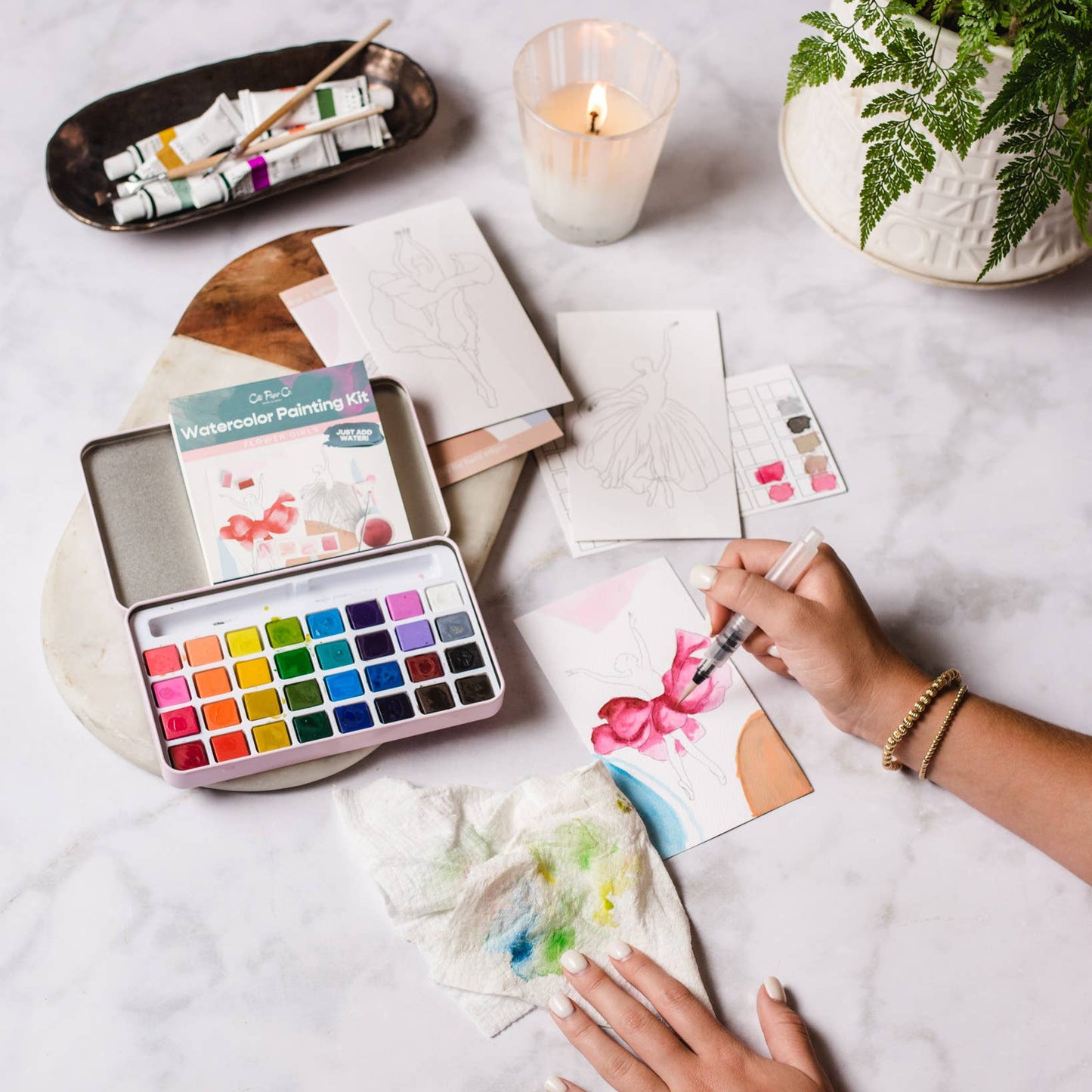 Person painting with watercolors on a table with art supplies and a candle.