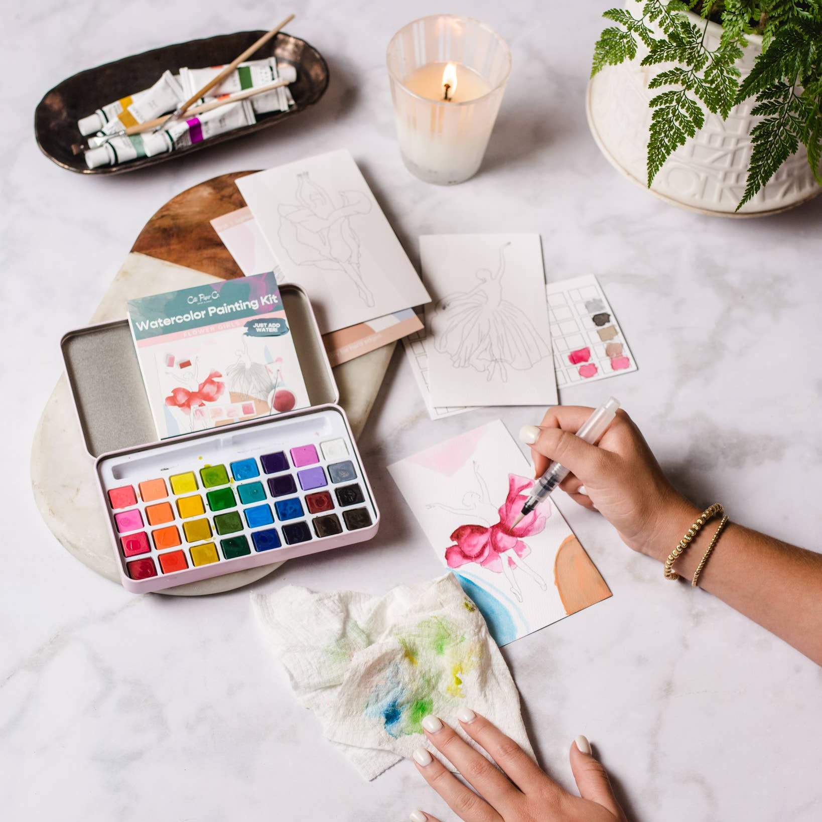 Person painting with watercolors on a table with art supplies and a candle.