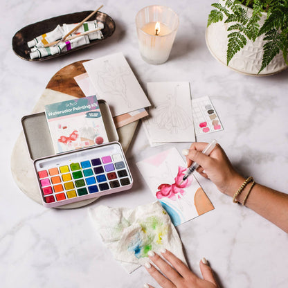 Person painting with watercolors on a table with art supplies and a candle.