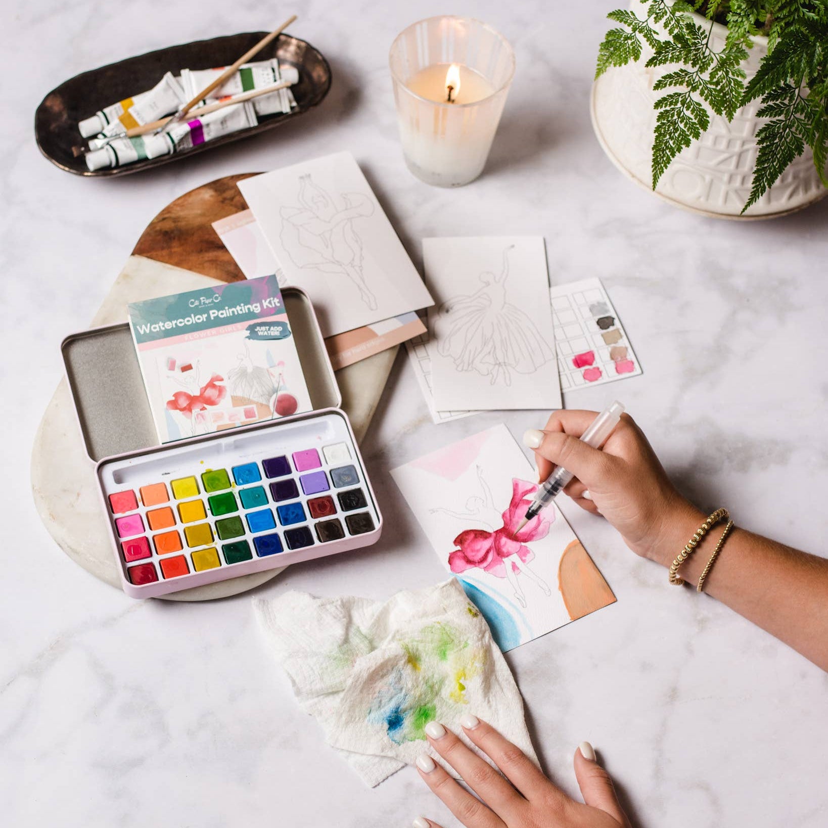 Person painting with watercolors on a table with art supplies and a candle.