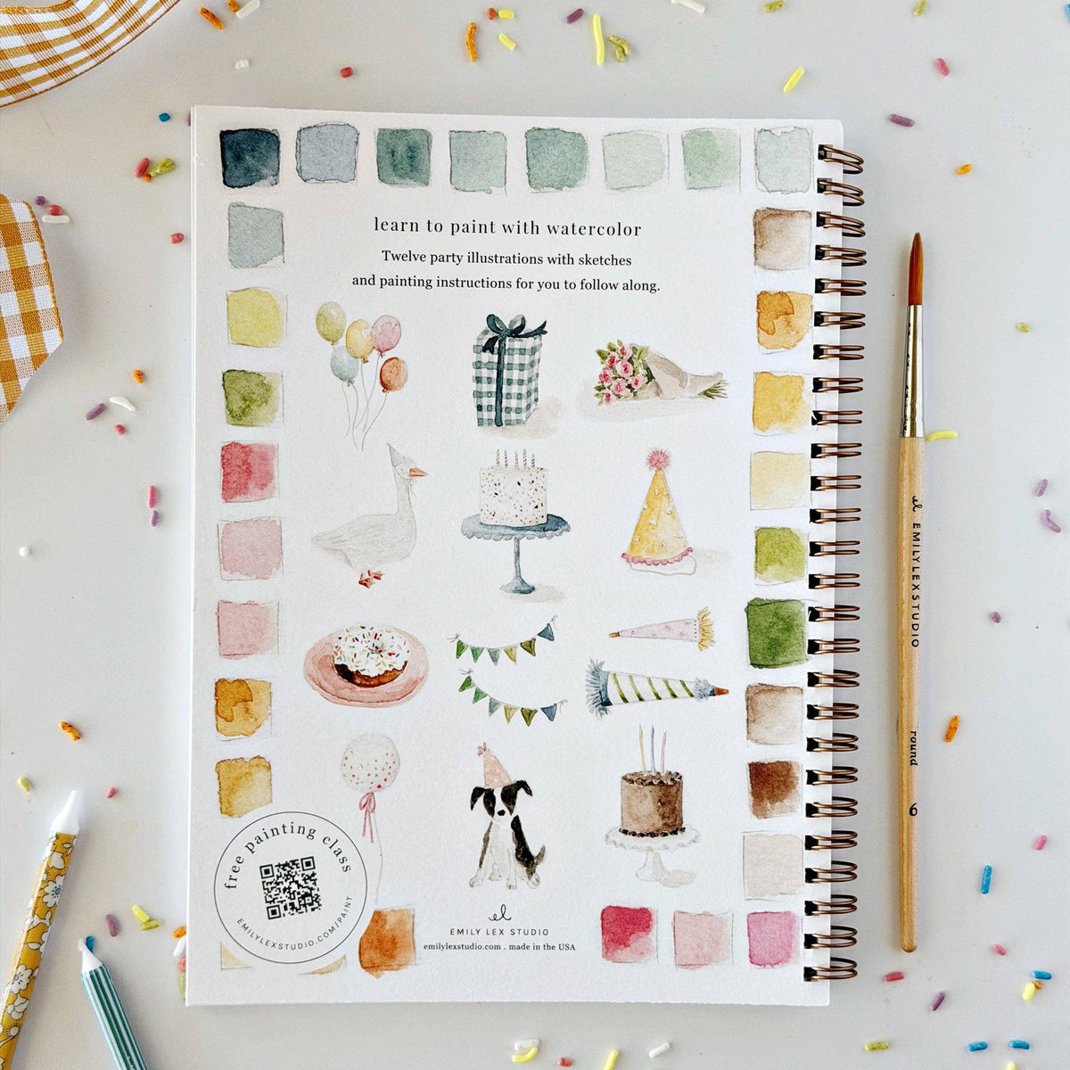 A spiral-bound watercolor painting guide book titled 'learn to paint with watercolor' is displayed on a light surface with colorful confetti. The book cover features various watercolor illustrations of birthday party themes like cakes, balloons, and hats, alongside a vertical column of watercolor swatches.