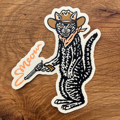 A die-cut vinyl sticker featuring a Western-style cat wearing a cowboy hat and bandana, holding a revolver, placed on a wooden surface. The sticker includes the word 'Swoon' in orange script.