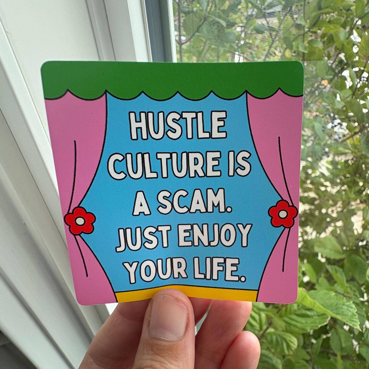 A hand holds a square printed card or sticker with a colorful cartoon curtain design and the text 'HUSTLE CULTURE IS A SCAM. JUST ENJOY YOUR LIFE.