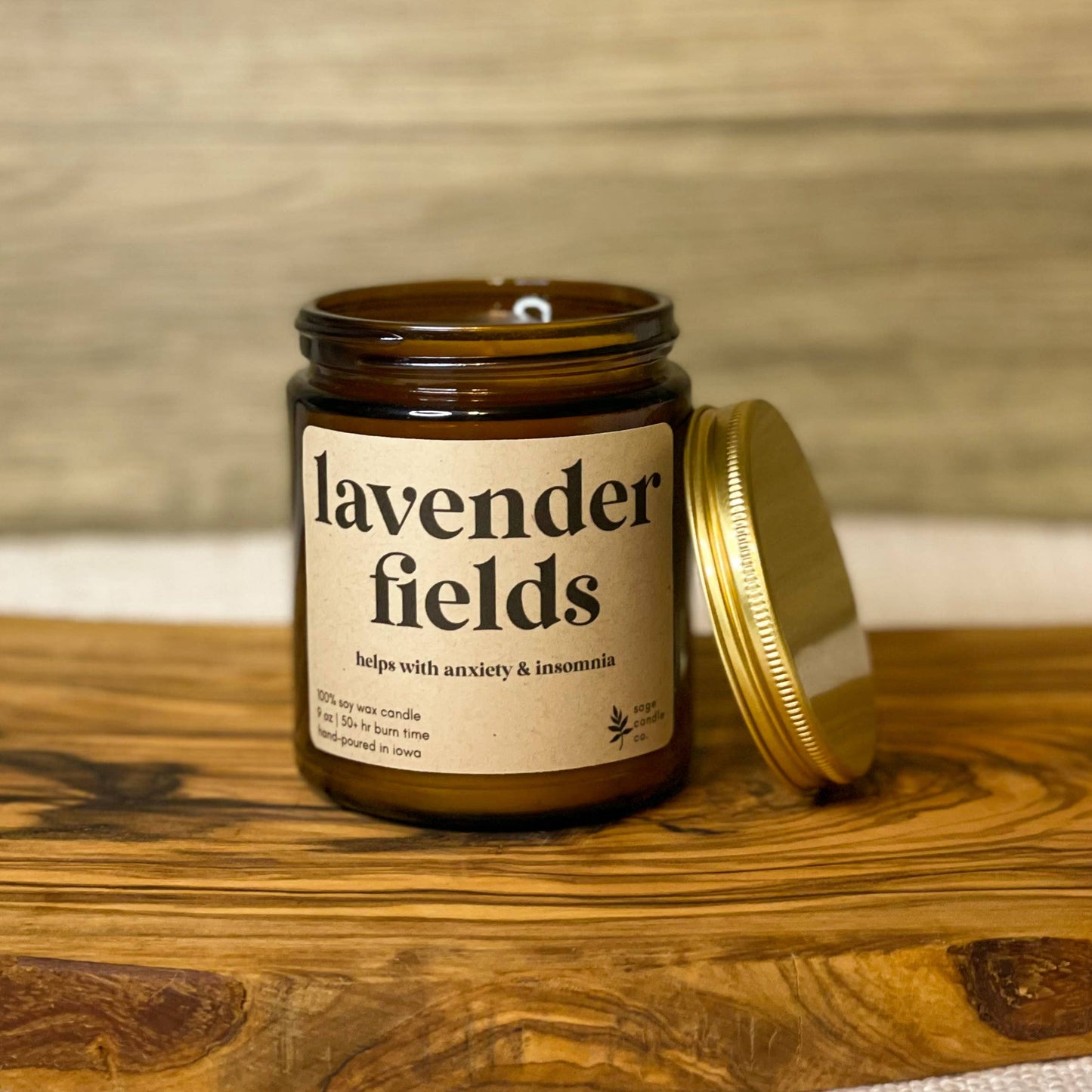 An amber glass jar candle labeled 'lavender fields' with a gold lid resting beside it on a rustic wooden board against a wood-paneled background.
