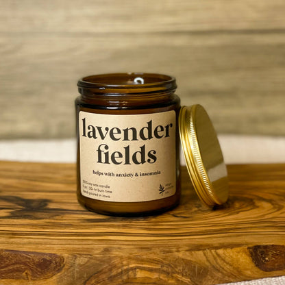 An amber glass jar candle labeled 'lavender fields' with a gold lid resting beside it on a rustic wooden board against a wood-paneled background.