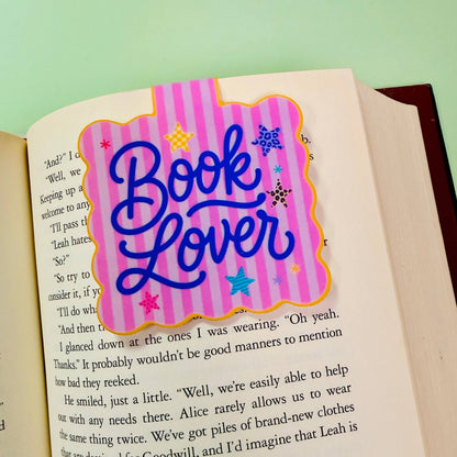 A decorative book lover bookmark with the words 'Book Lover' in blue script on a pink striped background, placed on an open book page.