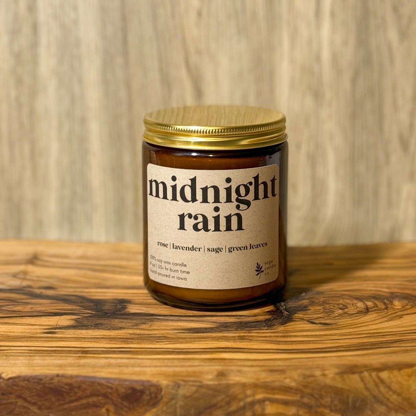 A candle jar labeled 'midnight rain' with a gold lid and amber glass, placed on a wooden board against a wood plank background.