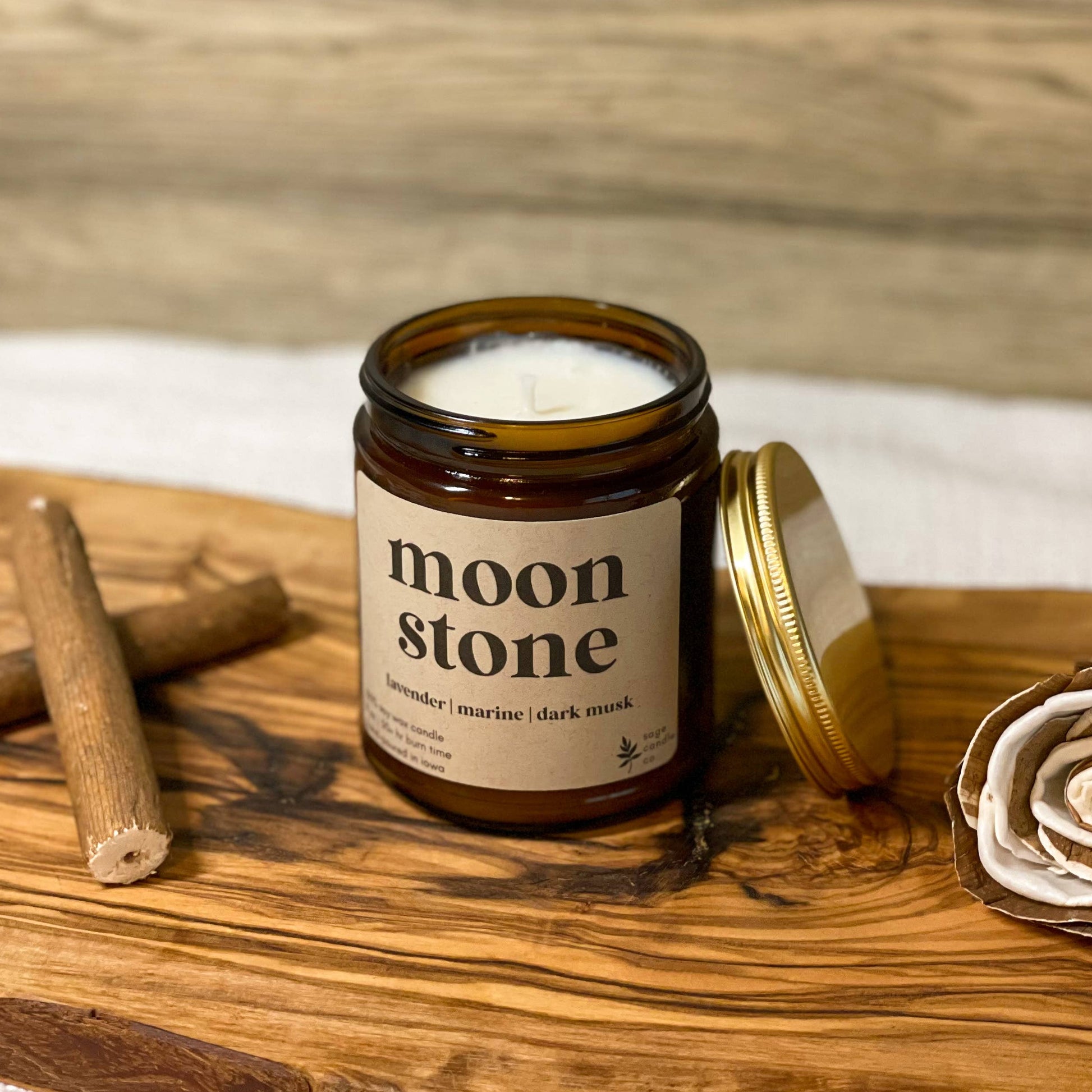 An amber glass jar candle with a cream label reading 'moon stone' sits on a rustic wooden board, with its gold lid leaning beside it. Two cinnamon sticks and a dried rose are placed around it, with a light wood plank background.