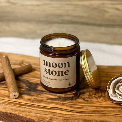 An amber glass jar candle with a cream label reading 'moon stone' sits on a rustic wooden board, with its gold lid leaning beside it. Two cinnamon sticks and a dried rose are placed around it, with a light wood plank background.