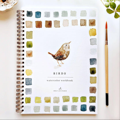 Watercolor workbook with bird illustration on a white surface with a cup and plant.