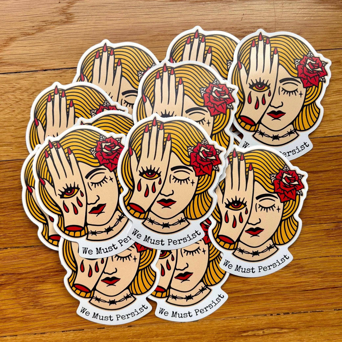 A set of die-cut stickers featuring a stylized illustration of a woman with blonde hair, a red rose, and crossed hands, with the text 'We Must Persist' on a wooden surface.