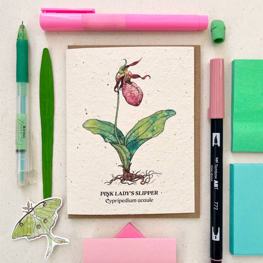 Card with pink lady's slipper illustration surrounded by stationery items on a light background