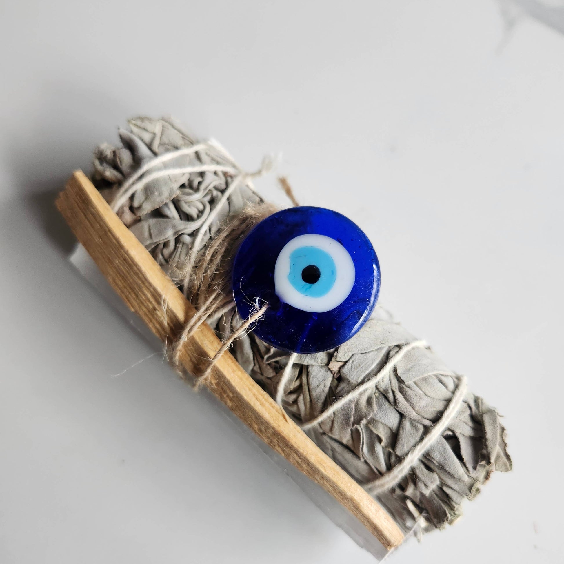 Incense stick wrapped with twine and featuring a blue evil eye charm on a light gray background
