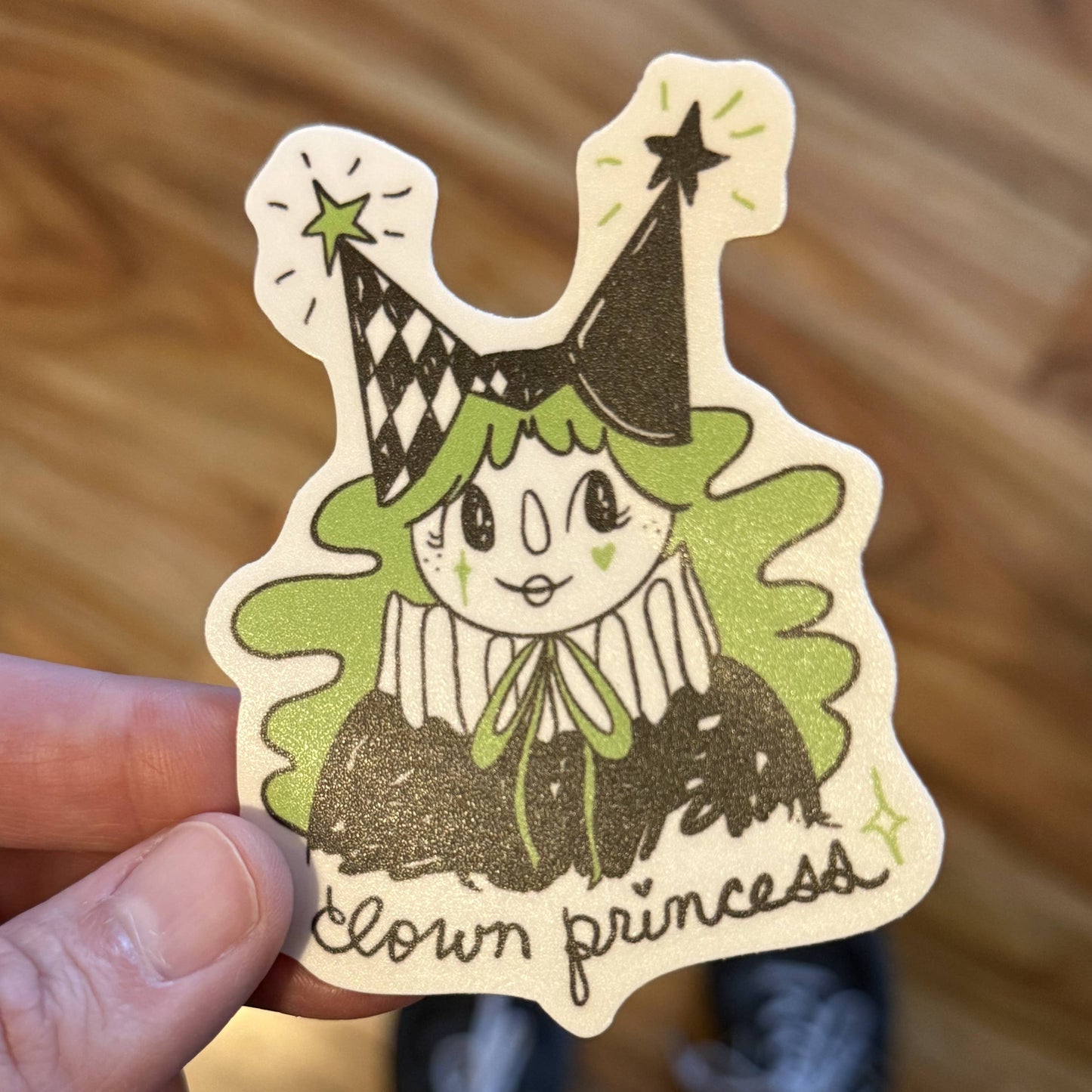 A hand holds a die-cut sticker featuring a cartoon clown princess with green hair, a black party hat, and the words 'clown princess' on a wooden floor background.