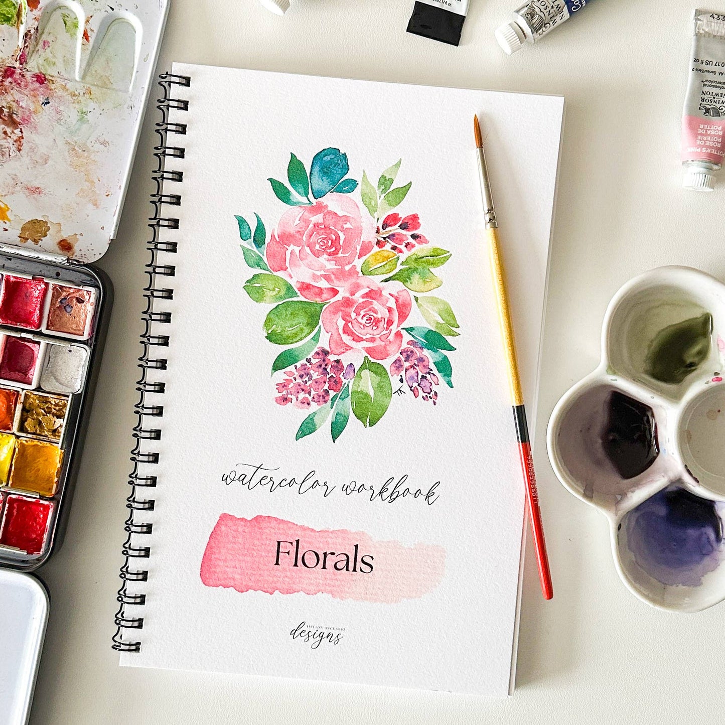 A spiral-bound watercolor workbook featuring a floral watercolor design on the cover, styled on a white surface with watercolor pans, a paintbrush, and paint tubes around it.