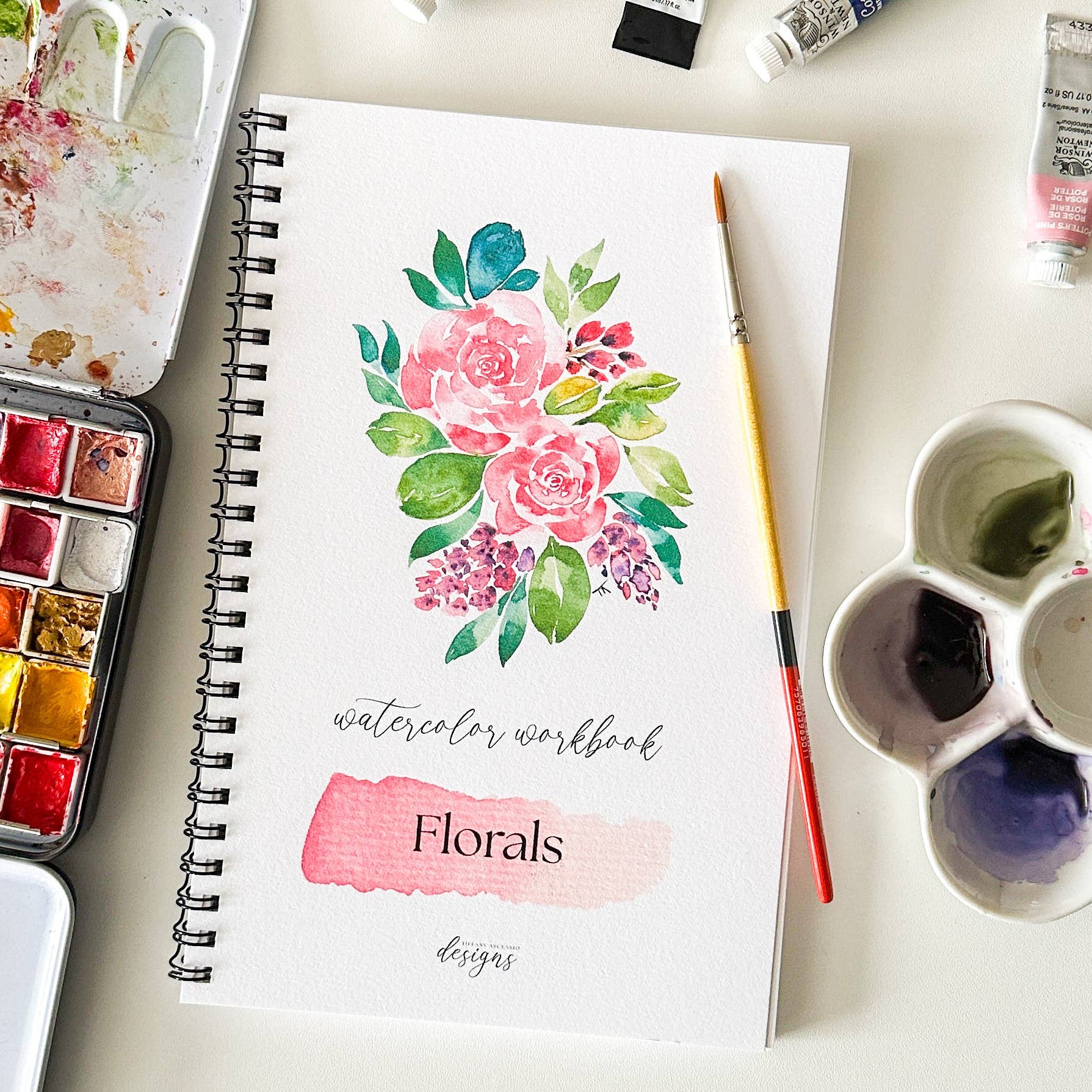 A spiral-bound watercolor workbook featuring a floral watercolor design on the cover, styled on a white surface with watercolor pans, a paintbrush, and paint tubes around it.