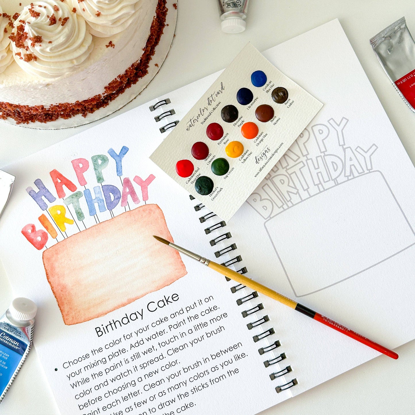Artistic birthday cake coloring book with watercolor paints and a cake on a table.