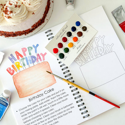 Artistic birthday cake coloring book with watercolor paints and a cake on a table.