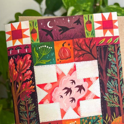 A hand holds a square art print or card featuring a colorful autumn quilt design with pumpkins, stars, trees, and leaves, set against a background of green houseplants.