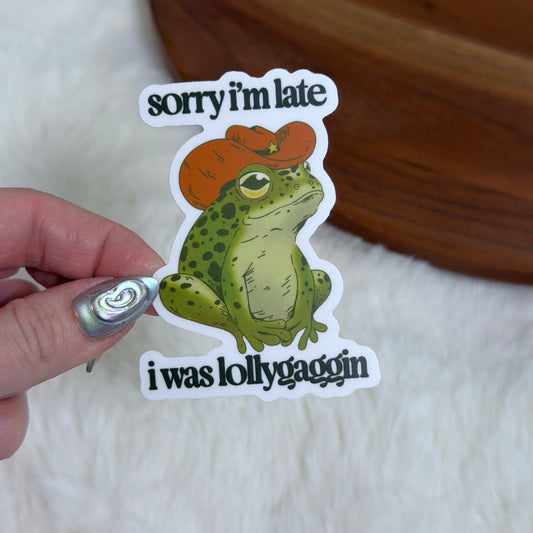 Sticker with a frog wearing a red hat and text held by a hand on a wooden surface.