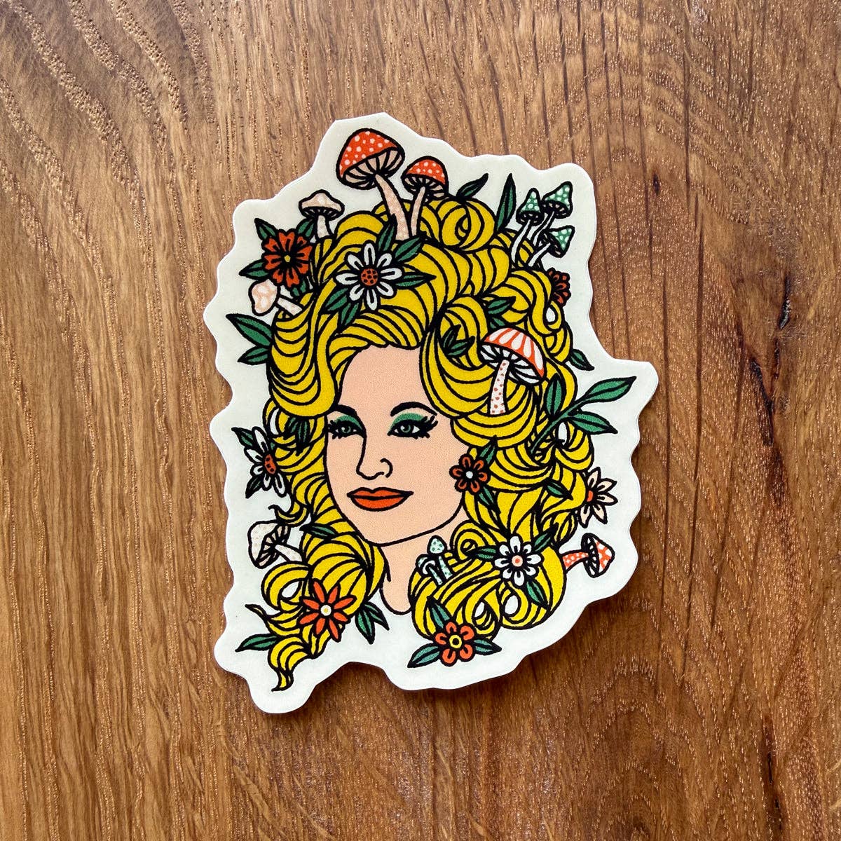 A die-cut sticker featuring an illustrated woman with voluminous yellow hair styled with mushrooms and flowers, placed on a brown wooden surface.