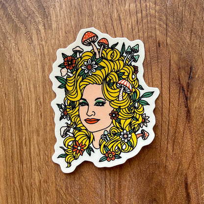 A die-cut sticker featuring an illustrated woman with voluminous yellow hair styled with mushrooms and flowers, placed on a brown wooden surface.