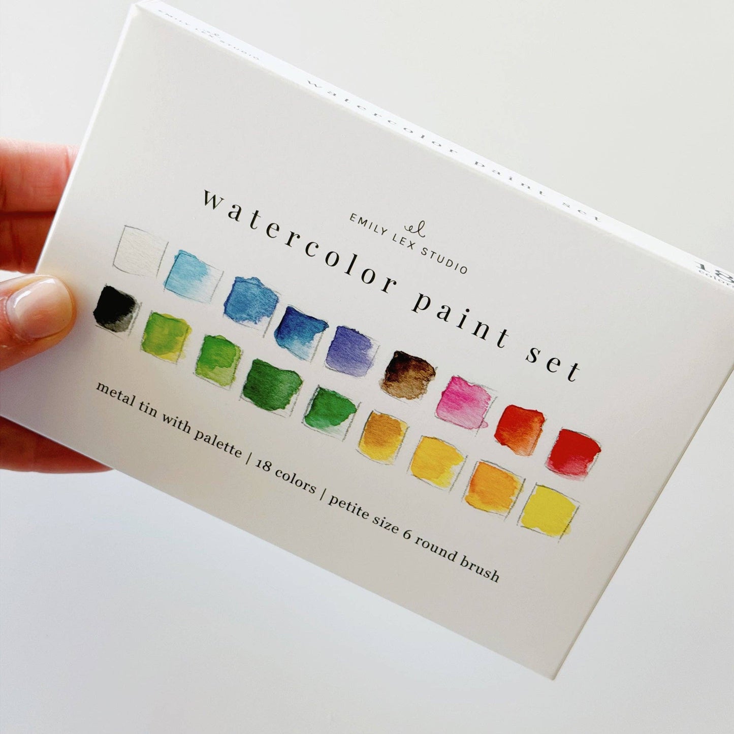 A hand holding a white watercolor paint set box by Emily Lee Studio. The box features printed watercolor swatches in multiple colors and text reading 'watercolor paint set' and 'metal tin with palette | 18 colors | petite size 6 round brush'.