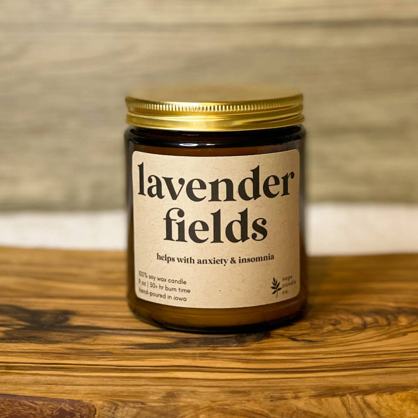 A candle jar labeled 'lavender fields' sits on a rustic wooden board against a light wood wall and white fabric background.