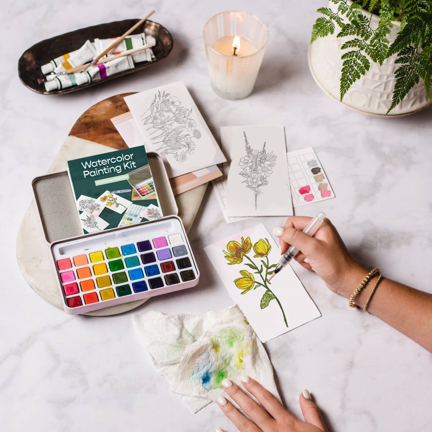 Person painting a flower with watercolors on a table with art supplies and a candle.