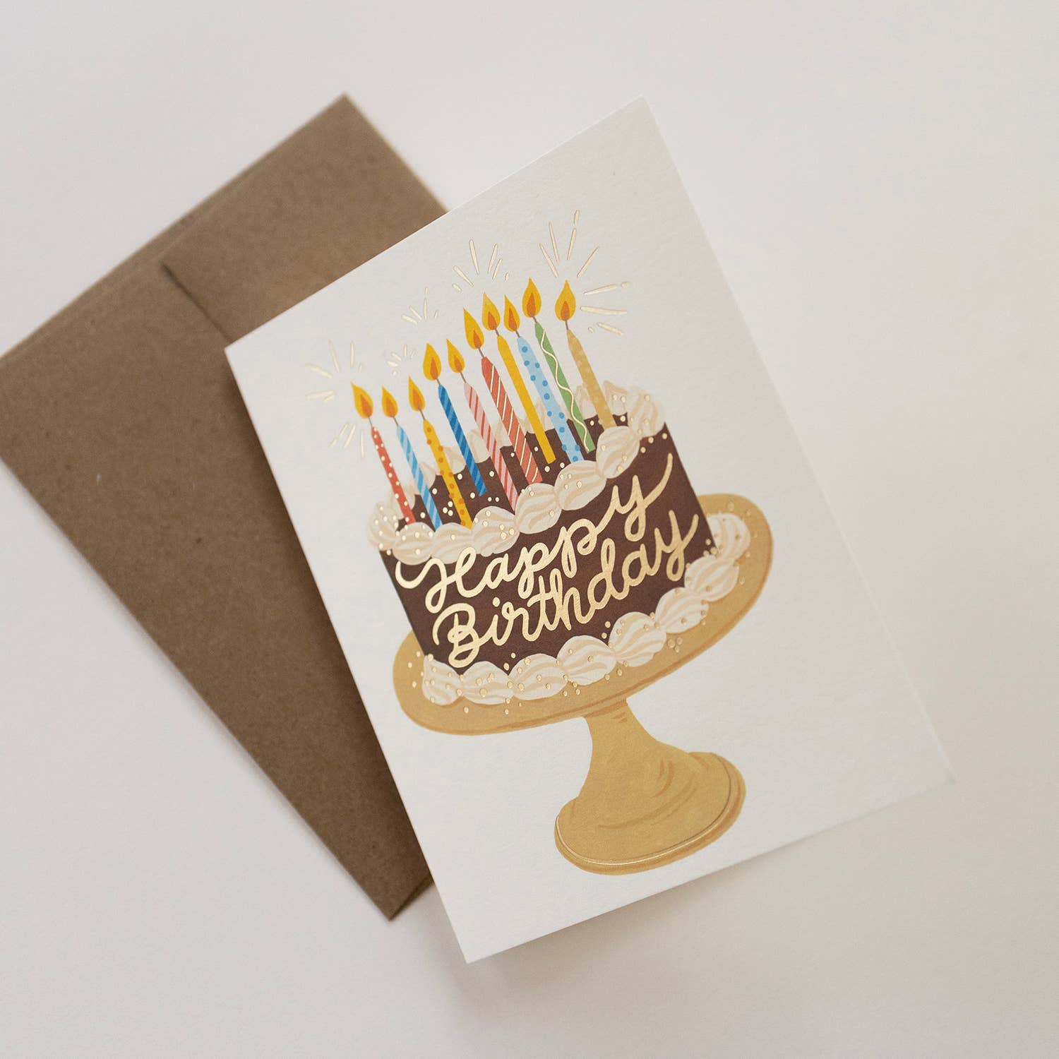 Birthday card with cake illustration and candles next to a brown envelope on a light background