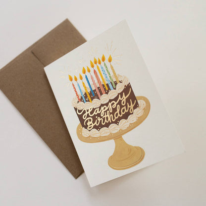 Birthday card with cake illustration and candles next to a brown envelope on a light background