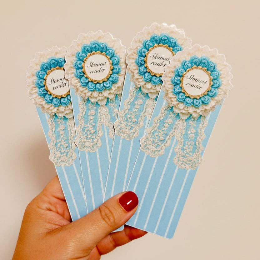 A hand with red nail polish holds a fan of five decorative book bookmarks. Each bookmark is light blue with white vertical stripes, featuring a white lace ribbon detail and a rosette-style embellishment with blue and white beads and a gold circular center.