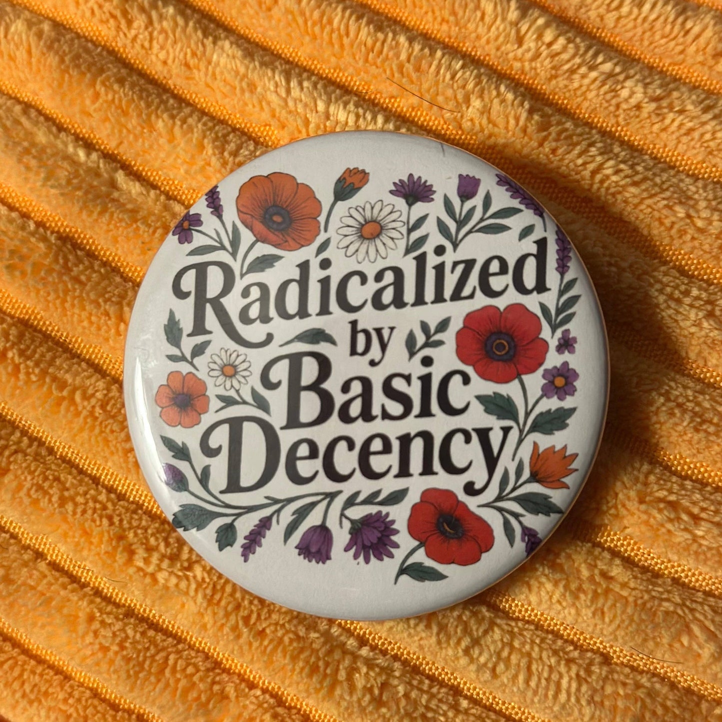 Button badge with floral design and text 'Radicalized by Basic Decency' on a textured orange background