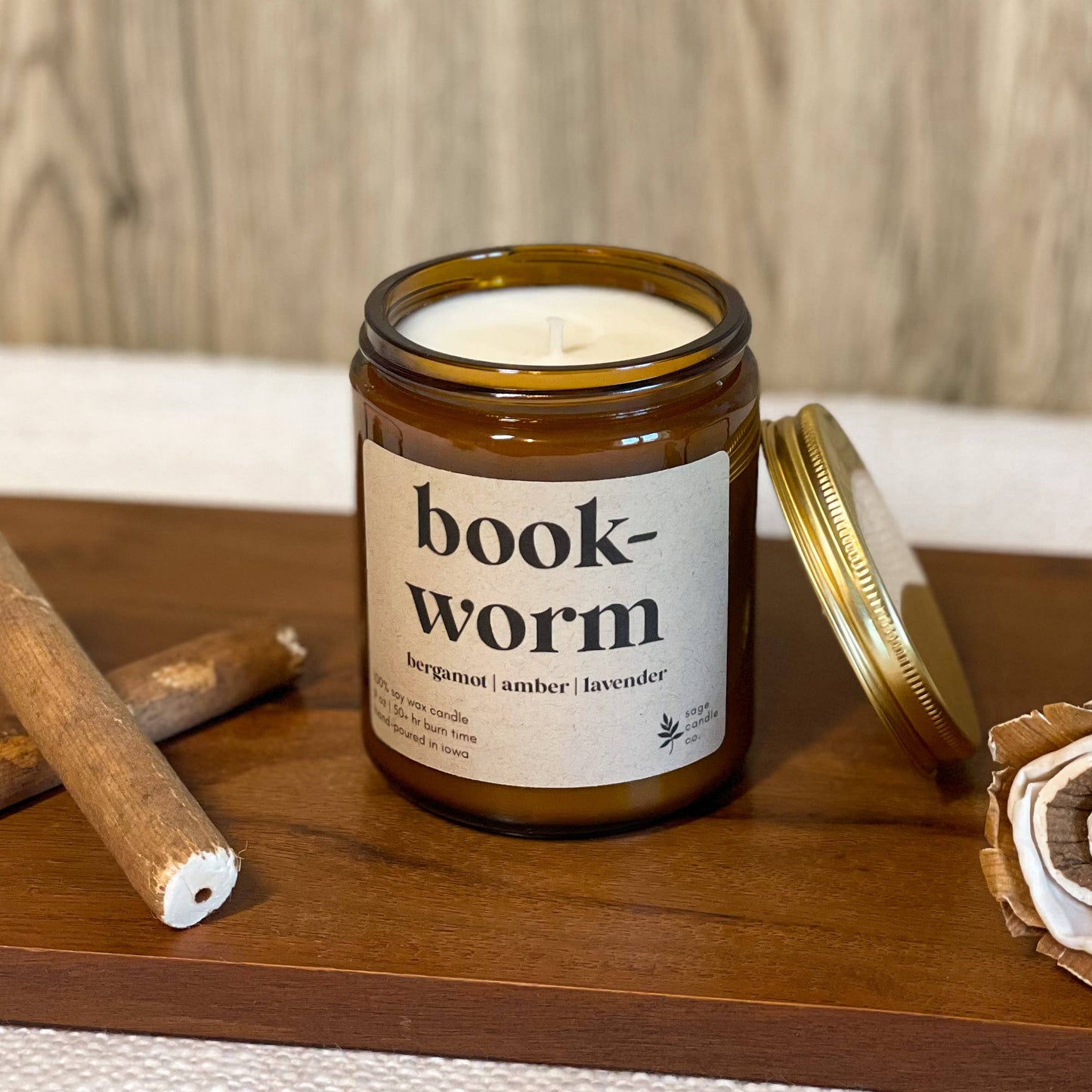 A candle in an amber glass jar with a white label reading 'book-worm' and a gold lid resting beside it on a wooden tray, styled with cinnamon sticks and a dried flower against a wood-grain background.
