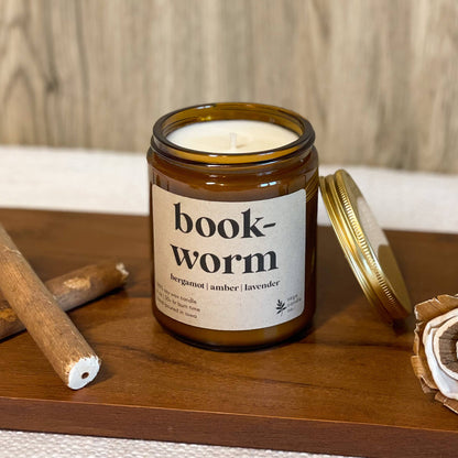 A candle in an amber glass jar with a white label reading 'book-worm' and a gold lid resting beside it on a wooden tray, styled with cinnamon sticks and a dried flower against a wood-grain background.