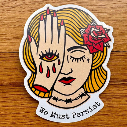 A die-cut vinyl sticker featuring a stylized illustration of a woman's face with one eye closed, red tears, a red rose in her hair, and the text 'We Must Persist' at the bottom, shown on a wooden surface.