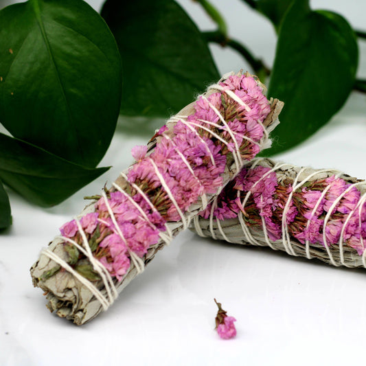 Two rolled sage bundles with pink flowers and white string on a white surface with green leaves in the background.