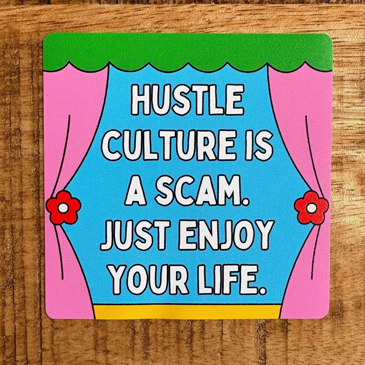 A square printed card or coaster with rounded corners sits on a wooden surface. The design shows pink curtains, a green top border, a yellow bottom band, and a bright blue center with large white text reading, 'HUSTLE CULTURE IS A SCAM.