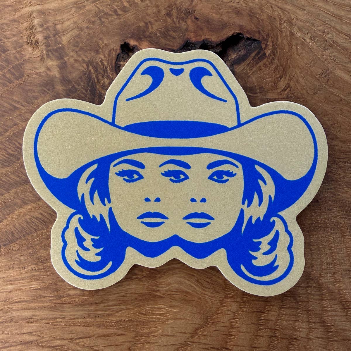A die-cut sticker or decal featuring a blue graphic of a woman's face wearing a cowboy hat, outlined in beige, placed on a textured wooden surface.