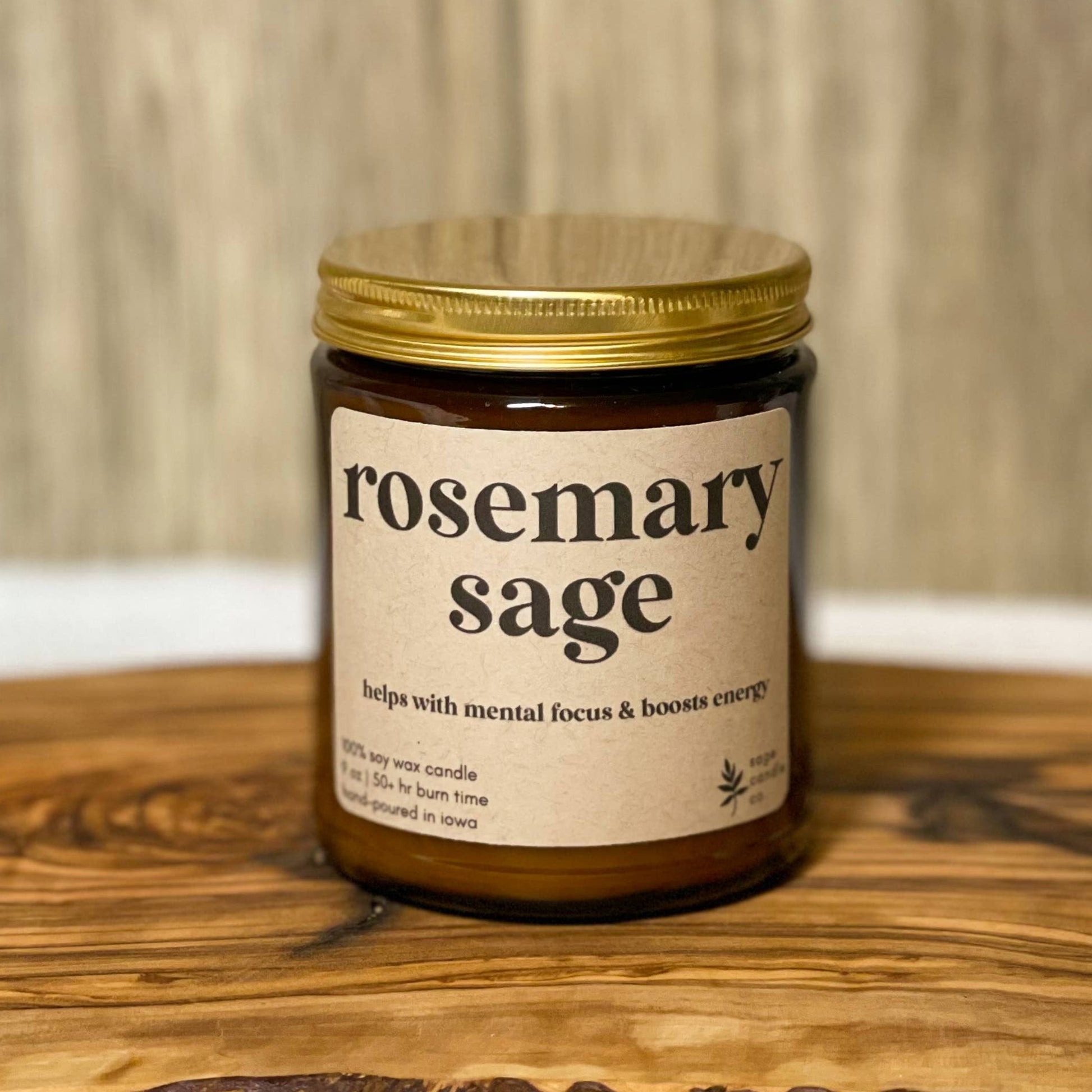 A single amber glass jar candle with a gold metal lid and a cream label reading 'rosemary sage' sits on a rustic live-edge wooden board against a light wood plank background.