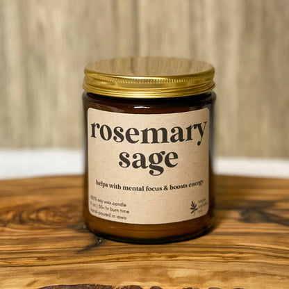 A single amber glass jar candle with a gold metal lid and a cream label reading 'rosemary sage' sits on a rustic live-edge wooden board against a light wood plank background.
