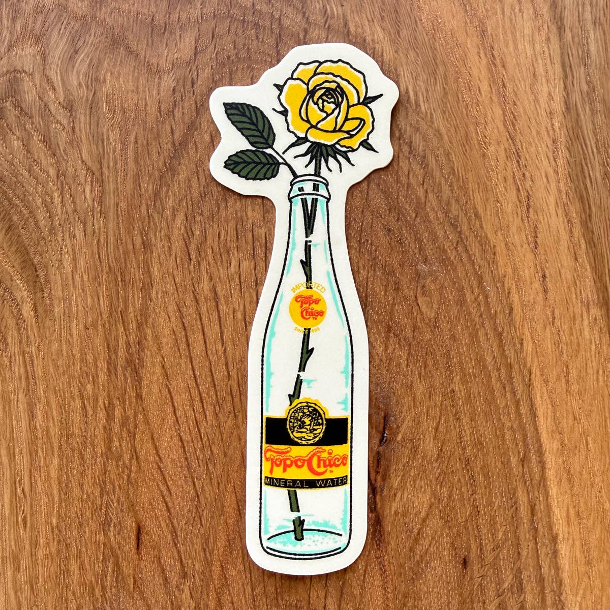 A die-cut sticker featuring a yellow rose in a clear glass bottle with a vintage Tojo Chico label, placed on a wooden surface.