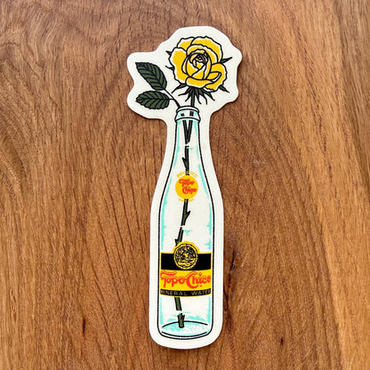 A die-cut sticker featuring a yellow rose in a clear glass bottle with a vintage Tojo Chico label, placed on a wooden surface.