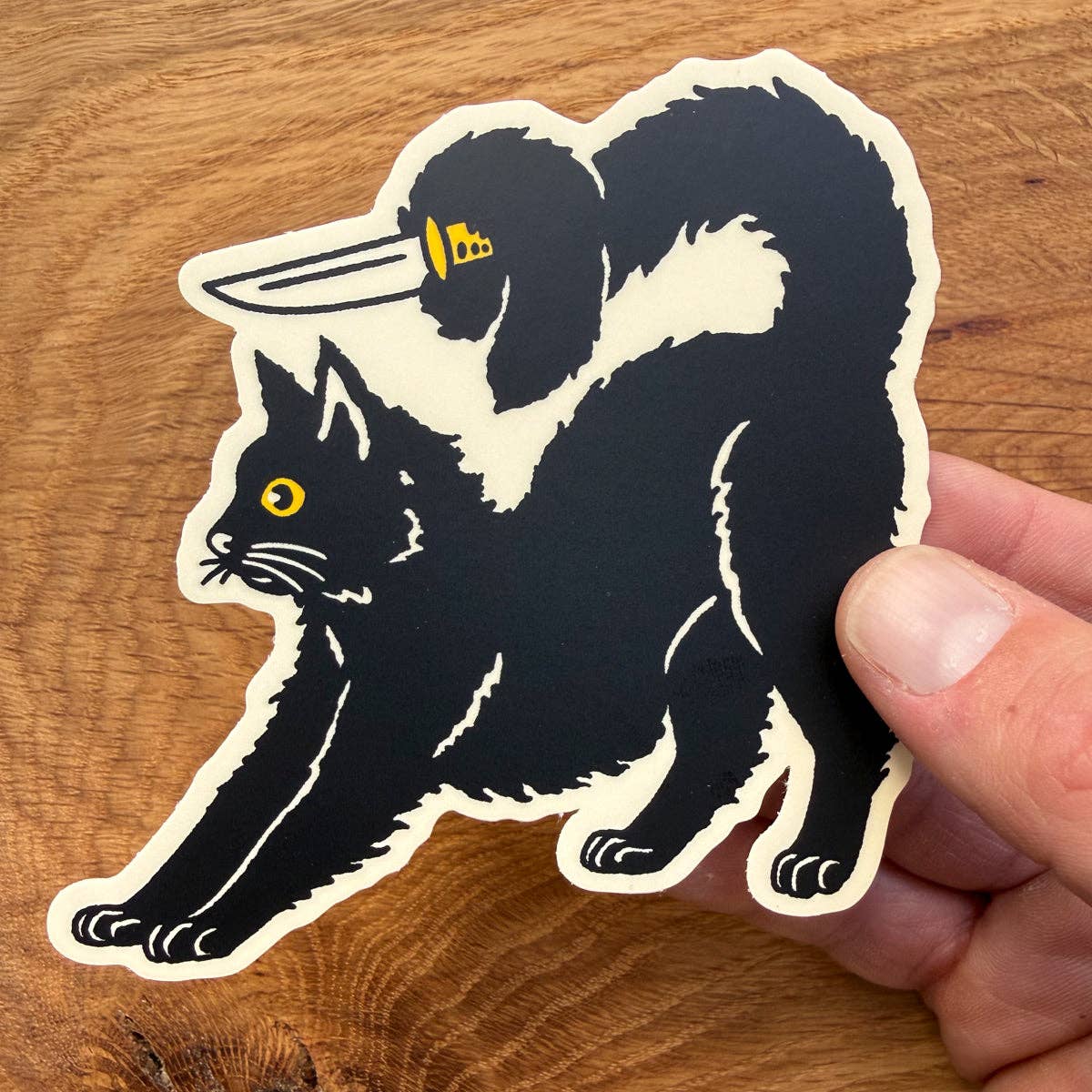 A die-cut sticker featuring a black cat with a yellow eye and a black dog carrying a knife on its back, held by a hand over a wooden surface.