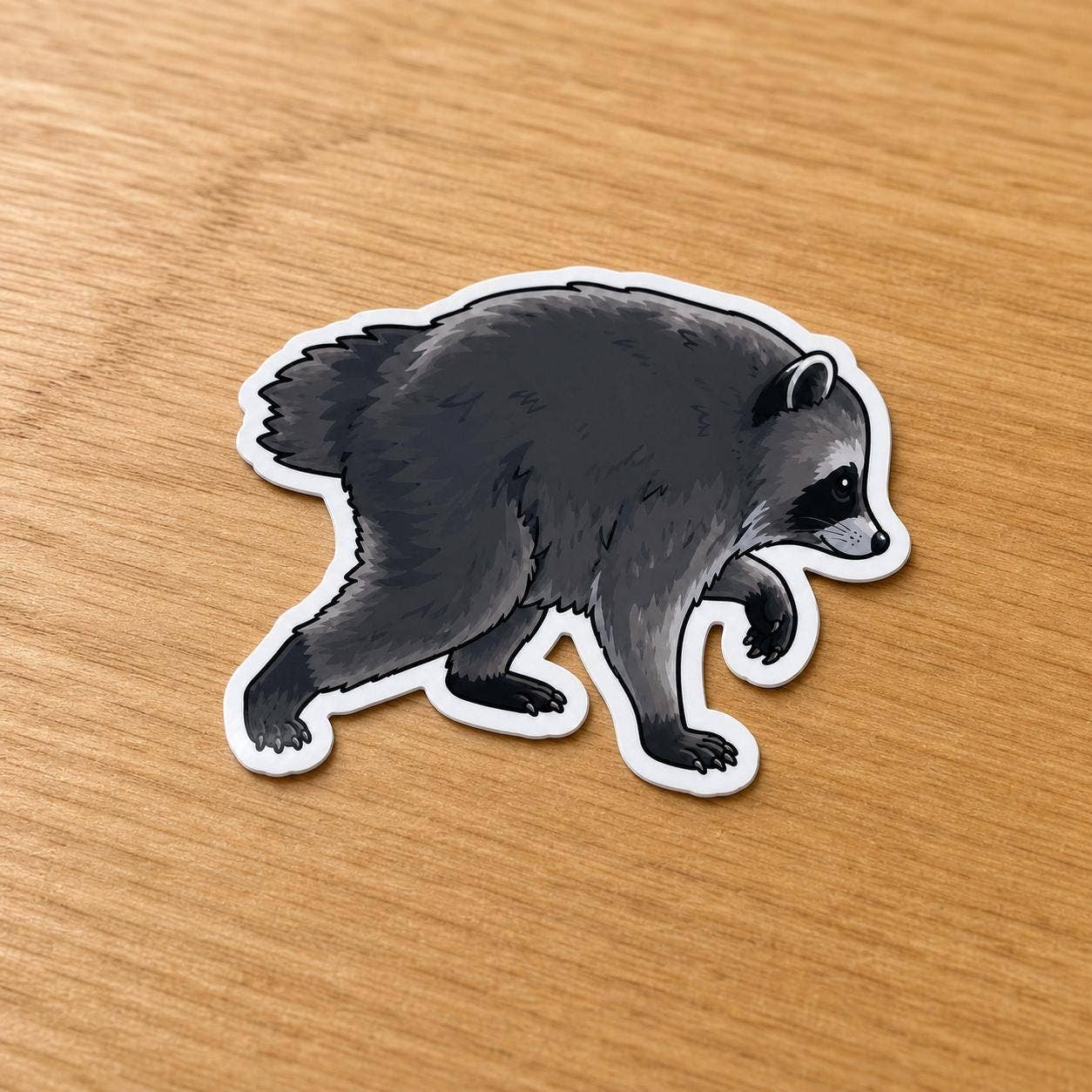 A die-cut vinyl sticker of a walking raccoon, shown on a light brown wooden tabletop. The raccoon is illustrated in dark gray and black with white facial markings and a white border outline.