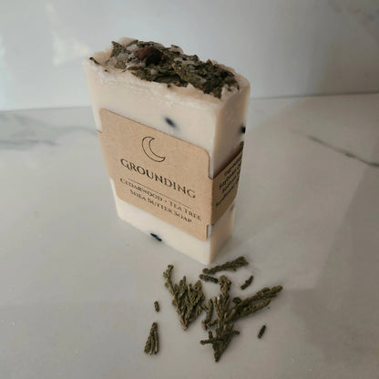 Bar of soap with a label on a white surface, surrounded by green herbs.